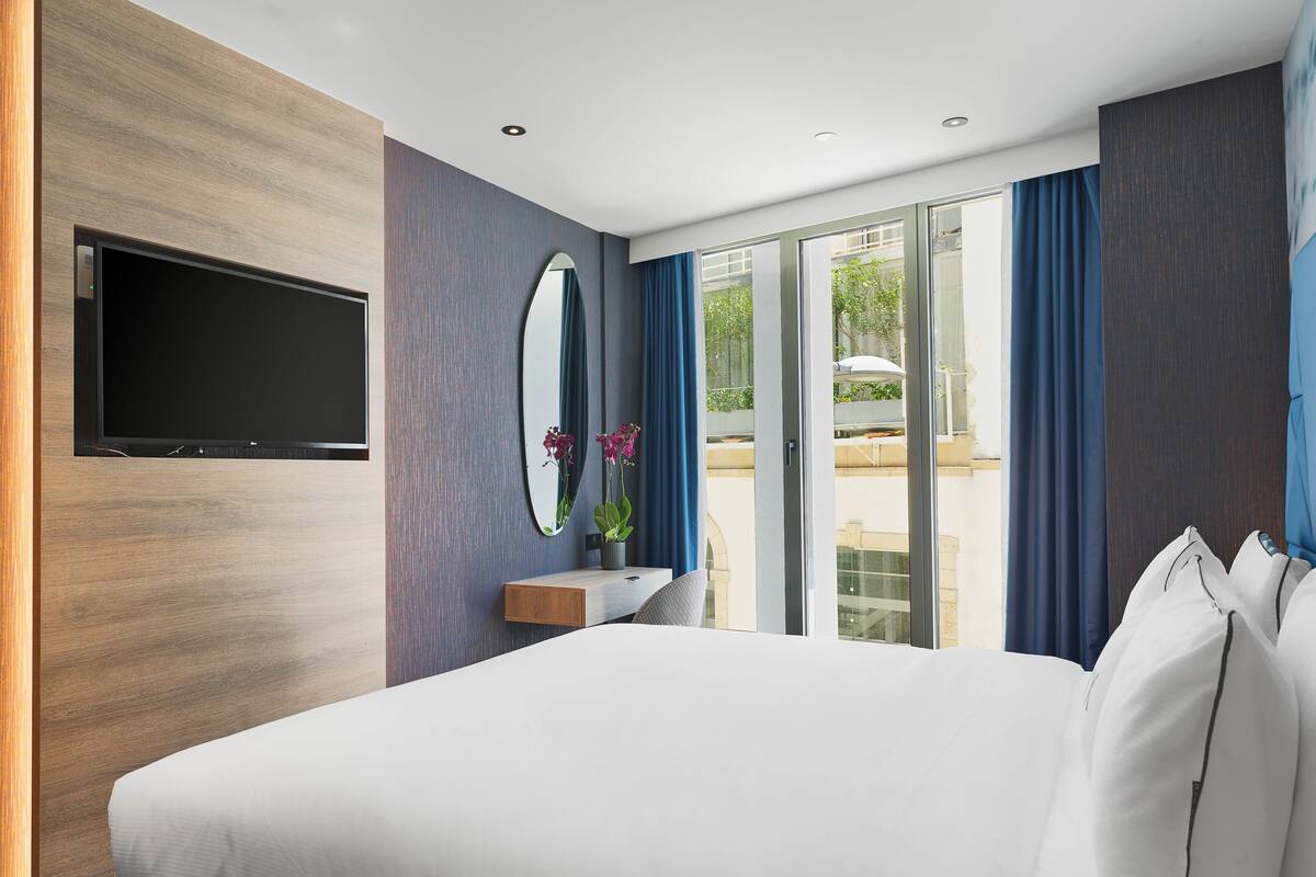 Una habitación de hotel moderna con una cama grande, un televisor de pantalla plana en la pared, una mesita con un florero y vistas al exterior a través de una puerta corrediza de vidrio.