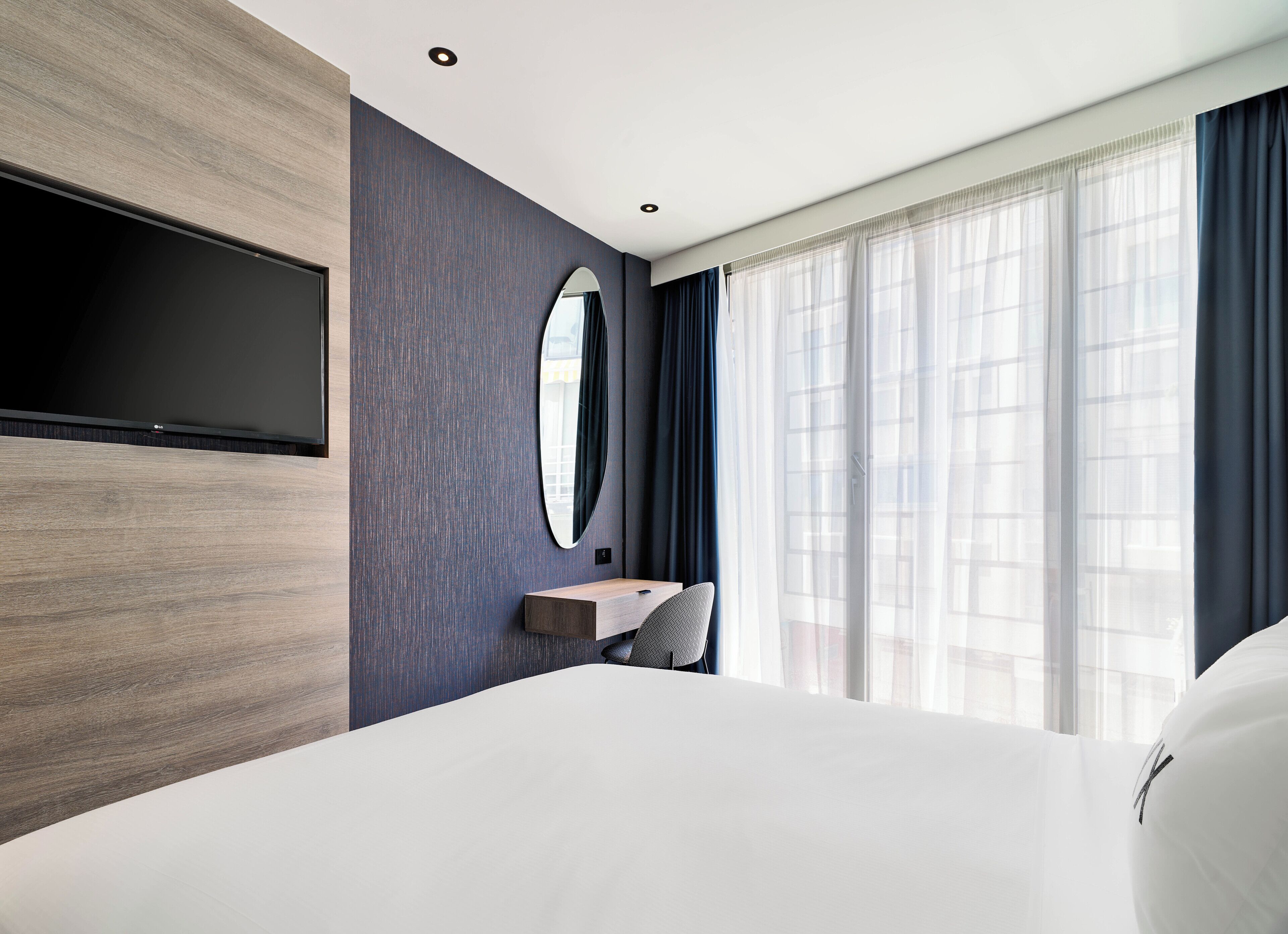 Una habitación de hotel moderna con una cama grande, un televisor de pantalla plana montado en la pared, un pequeño escritorio con una silla y un amplio ventanal con cortinas de gasa.