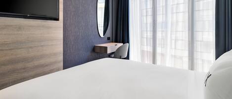 Una habitación de hotel moderna con una cama grande, un televisor de pantalla plana montado en la pared, un pequeño escritorio con una silla y un amplio ventanal con cortinas de gasa.