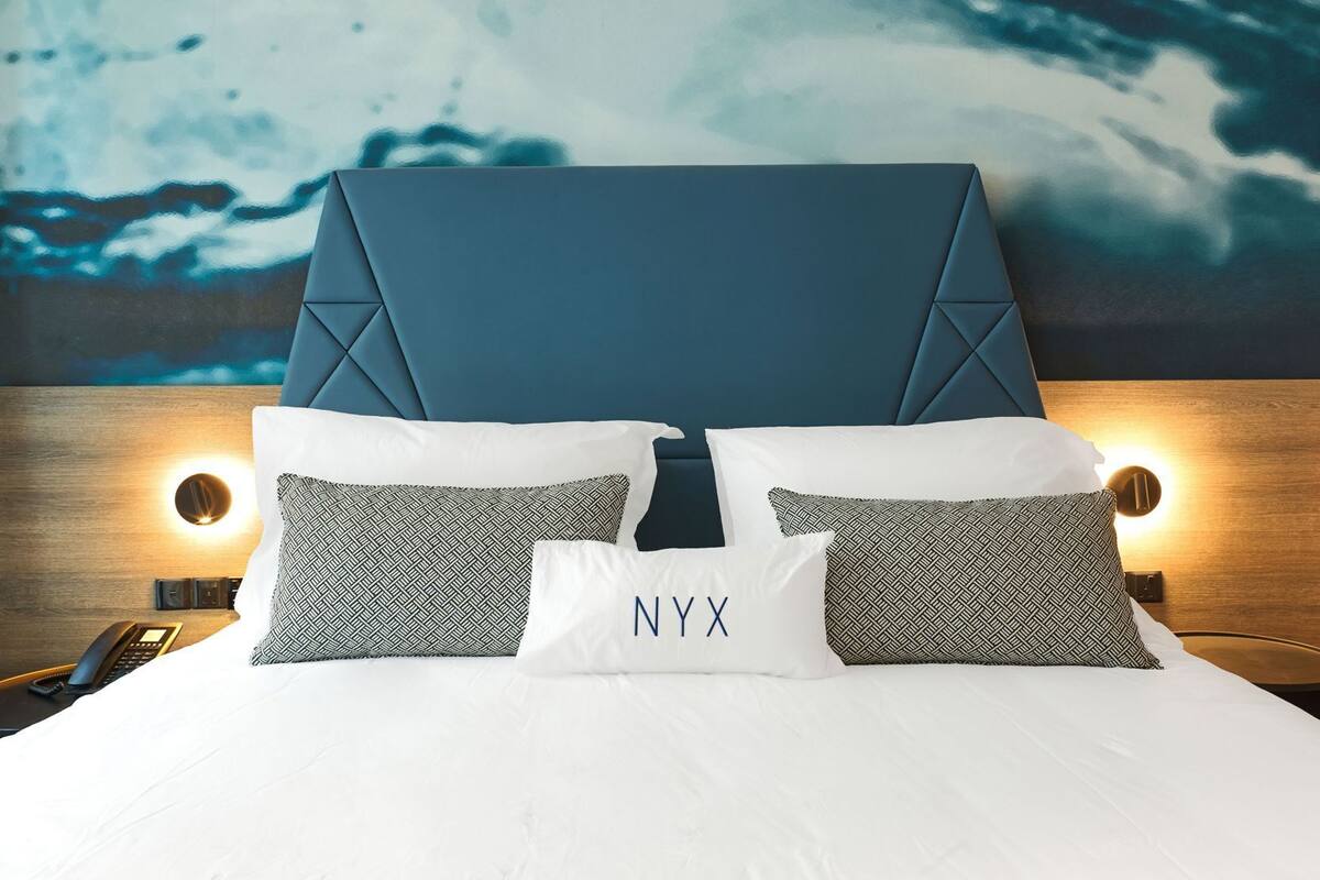 Una habitación de hotel con una cama grande, cabecera, dos almohadas estampadas, una almohada blanca con el texto 'NYX', lámparas de noche y un teléfono en la mesita de noche.