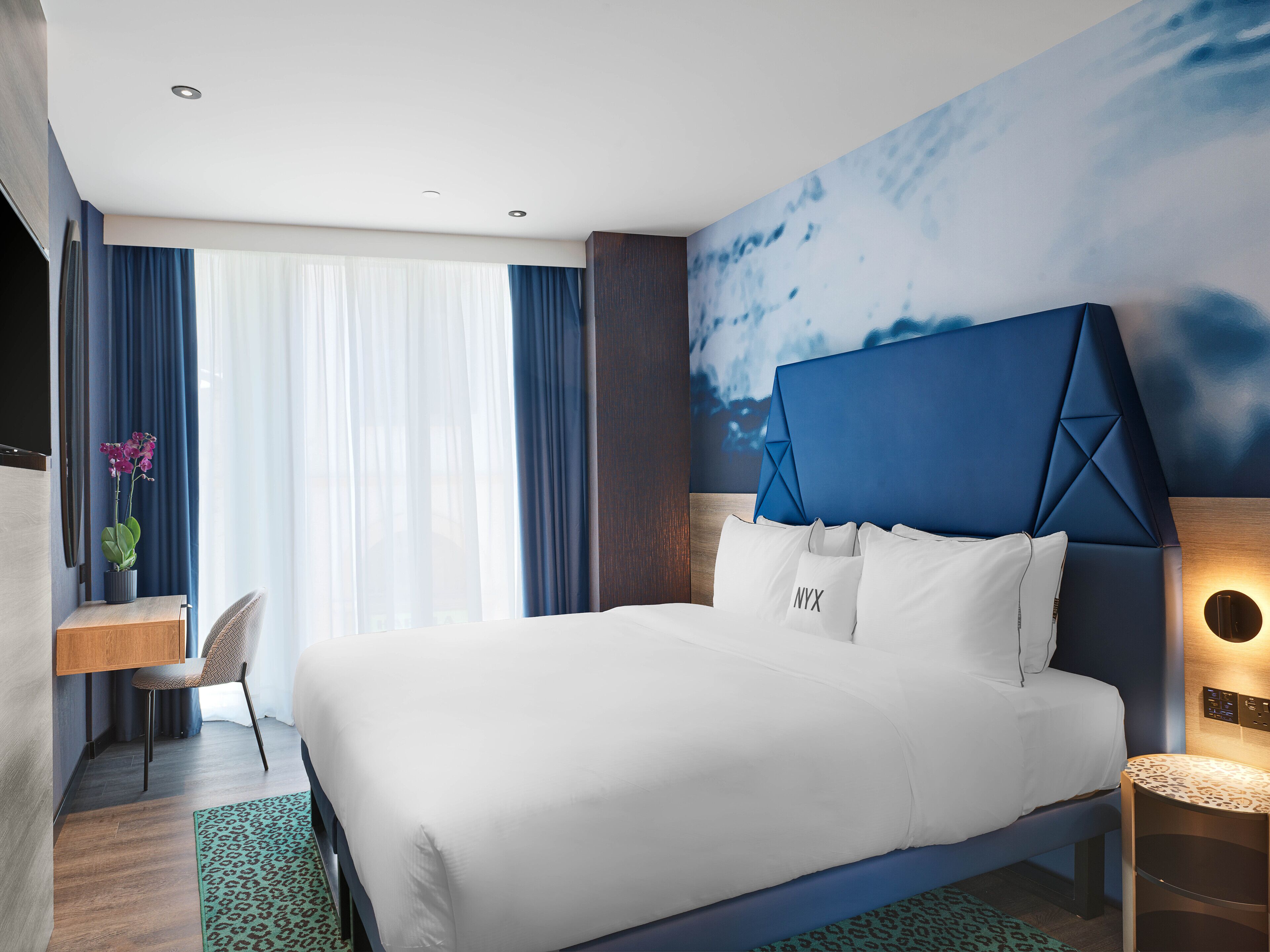 A modern hotel room with a large bed, a desk, and a chair. There is a window with curtains, a wall with a painting, and a nightstand with a lamp.