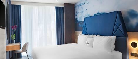 A modern hotel room with a large bed, a desk, and a chair. There is a window with curtains, a wall with a painting, and a nightstand with a lamp.