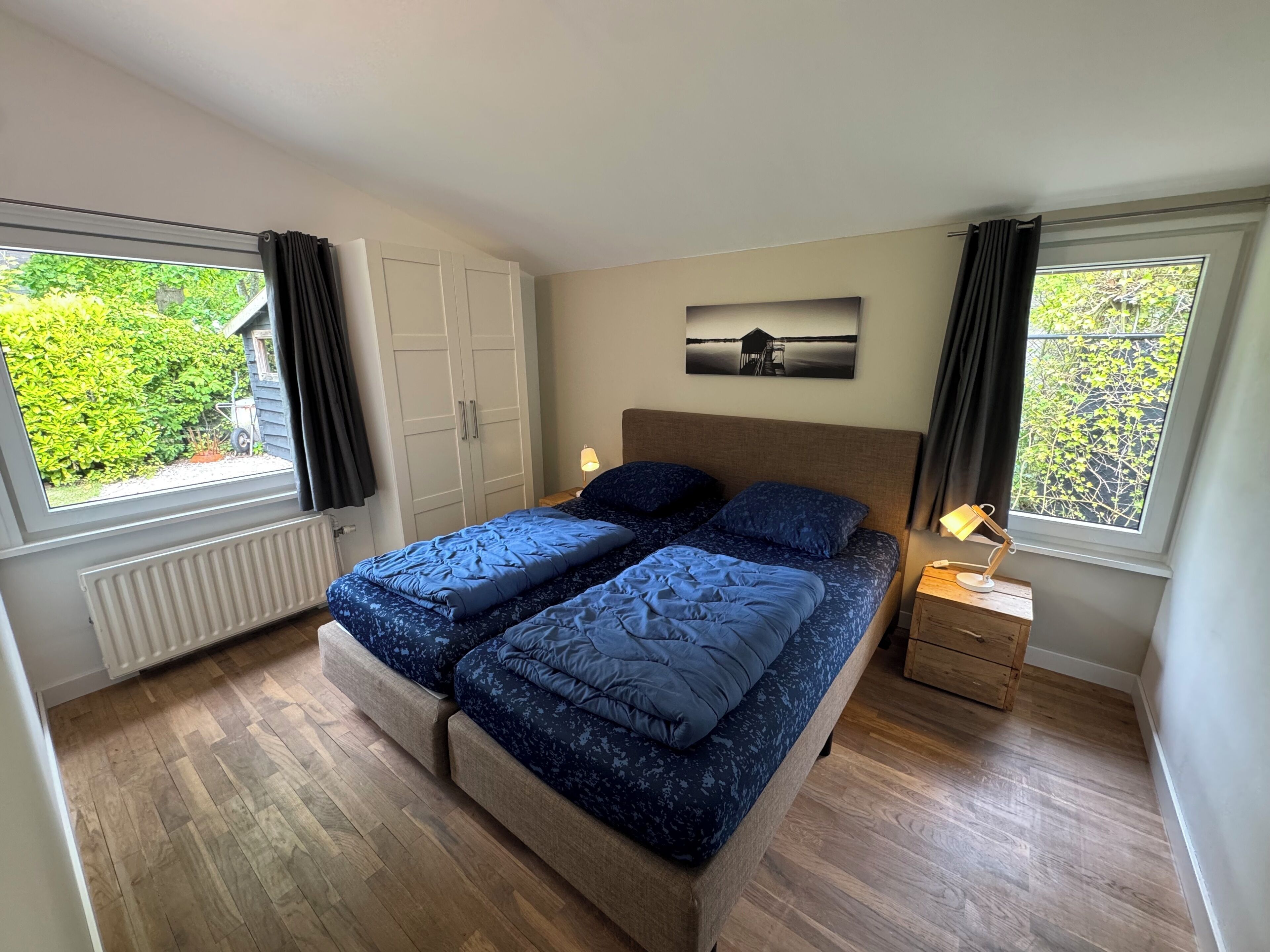 A bedroom with a double bed, two bedside tables, a radiator, and a window with a view of greenery.