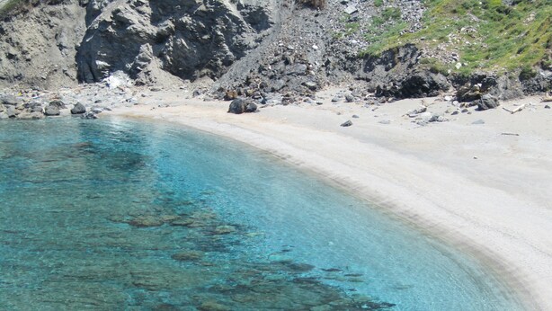 A clear turquoise beach with rocky formations and a rugged cliff in the background.