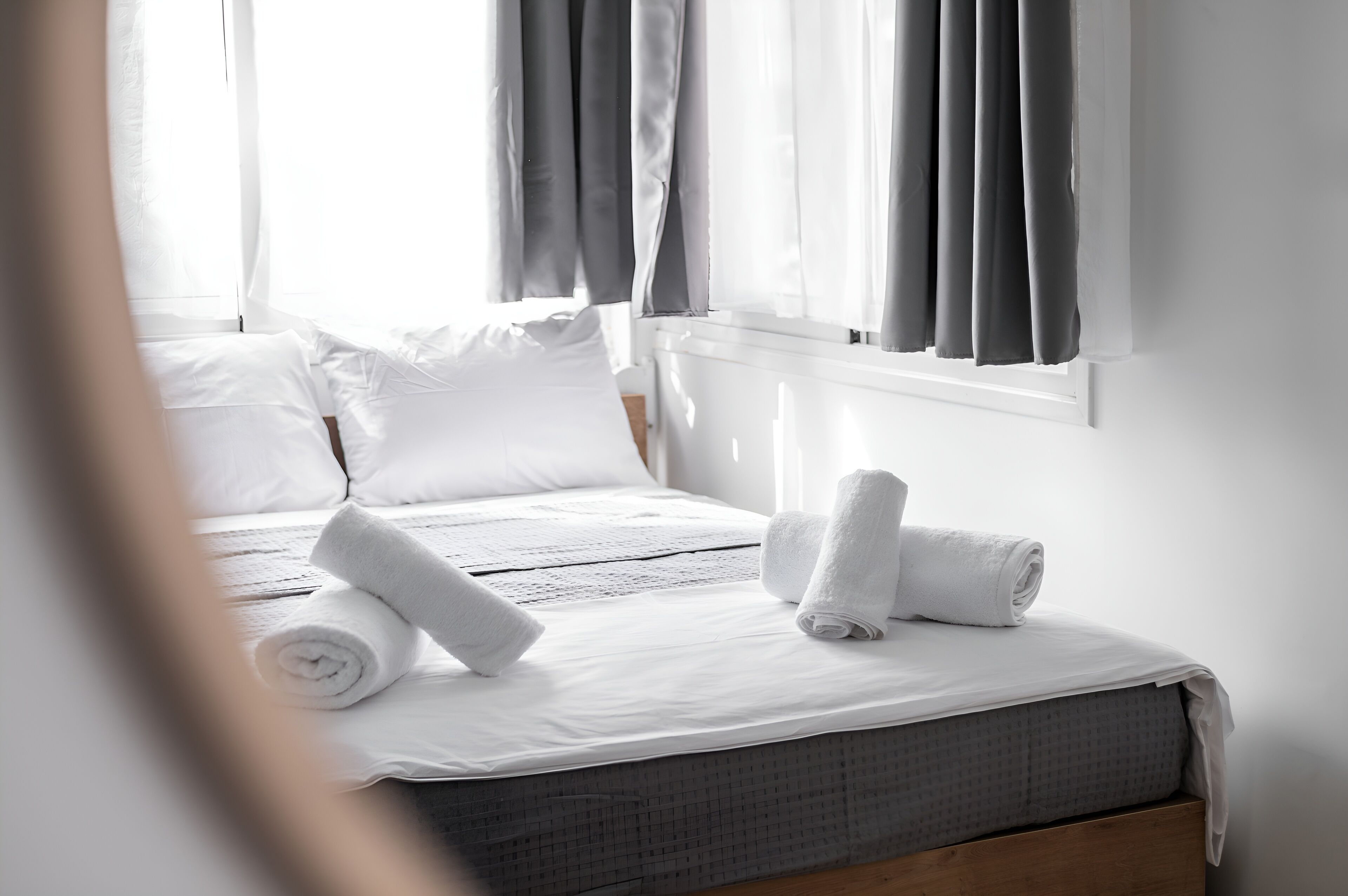 A neatly made bed with white linens and rolled towels, a window with grey curtains, and a white wall.