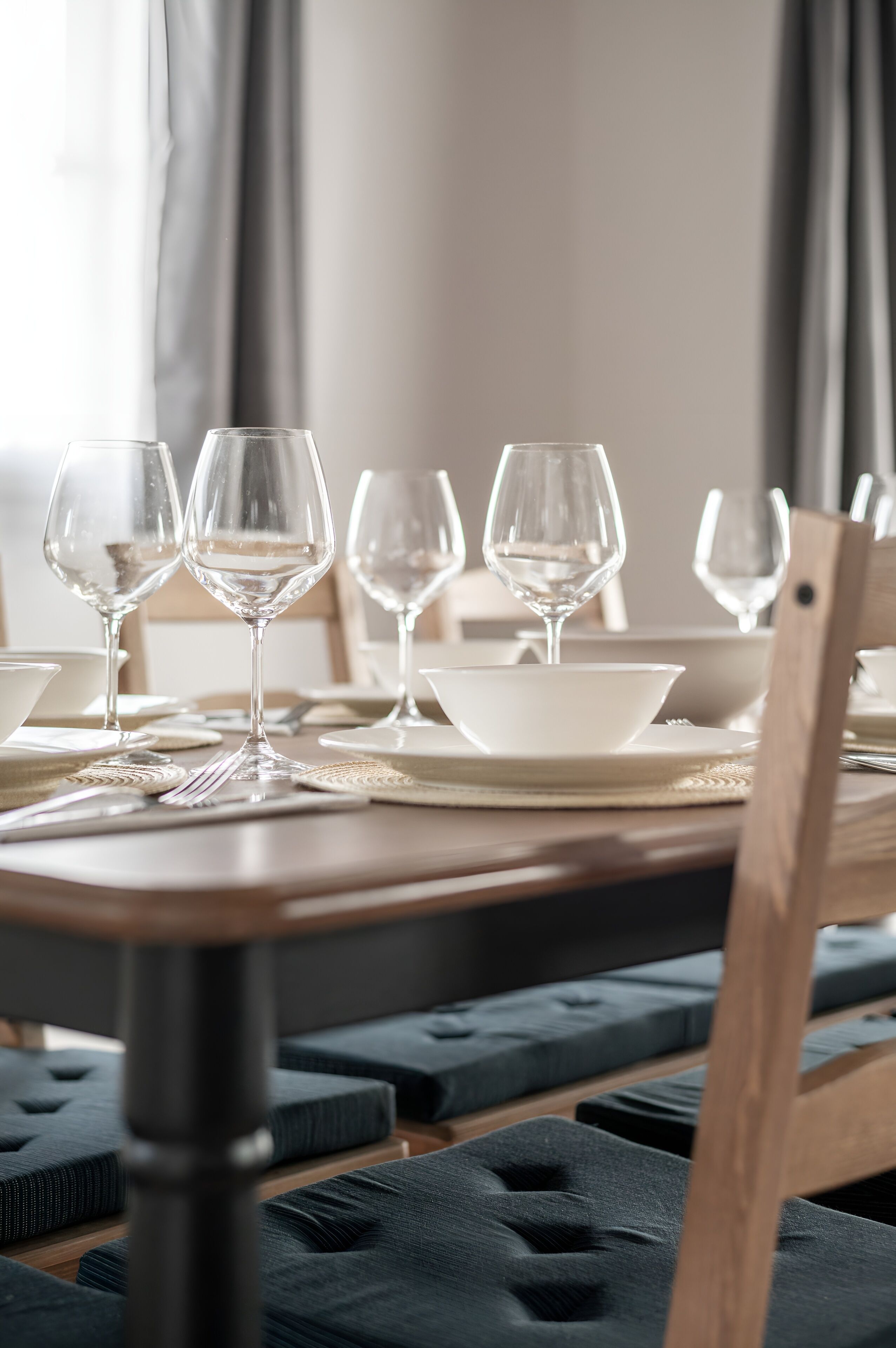 A dining table set with white plates, bowls, and wine glasses, with dark cushioned chairs around it.