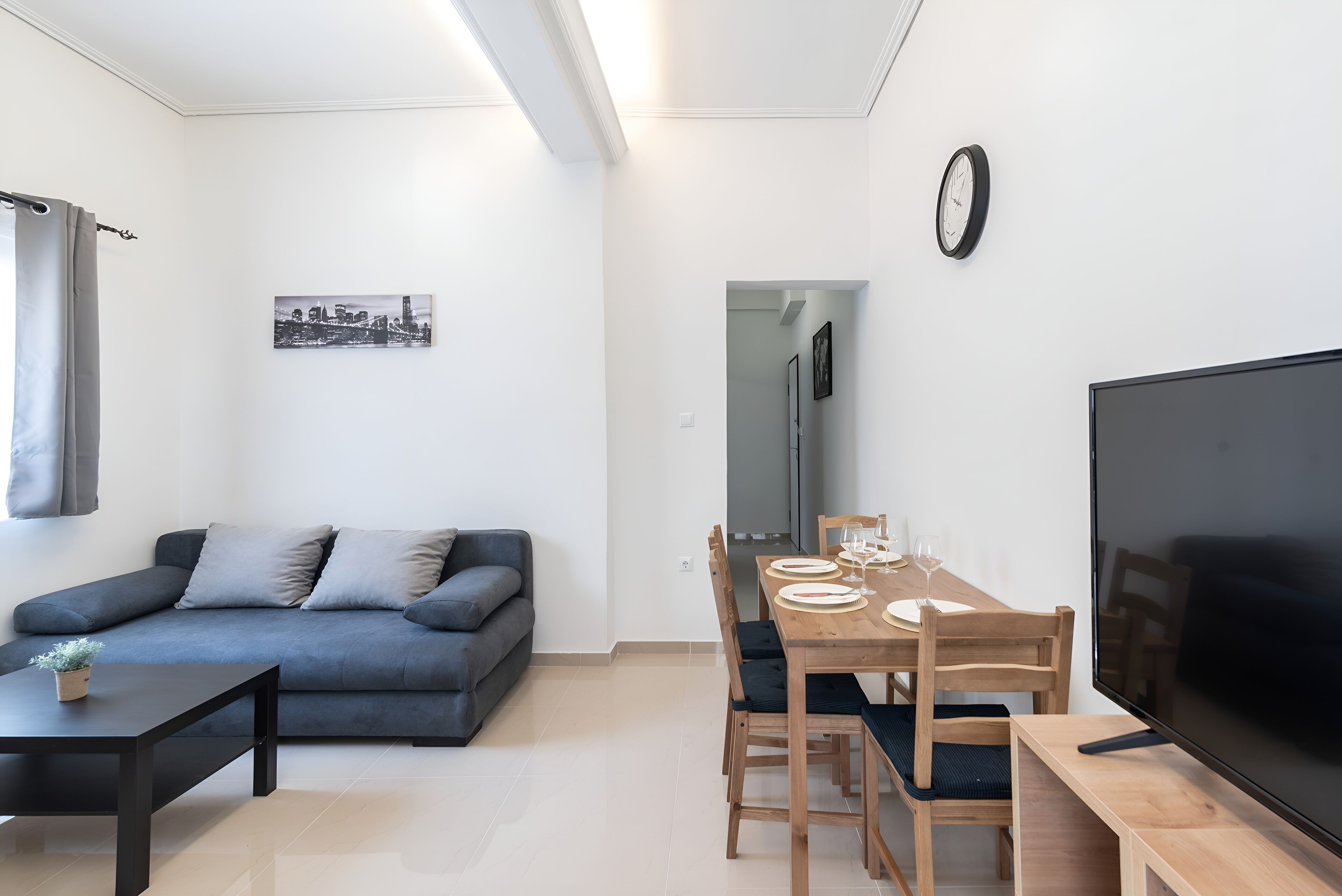 Ein modernes Wohnzimmer mit einem grauen Sofa, einem Holzesstisch gedeckt mit Tellern und Gläsern sowie einem Flachbildfernseher auf einem Holzschrank.