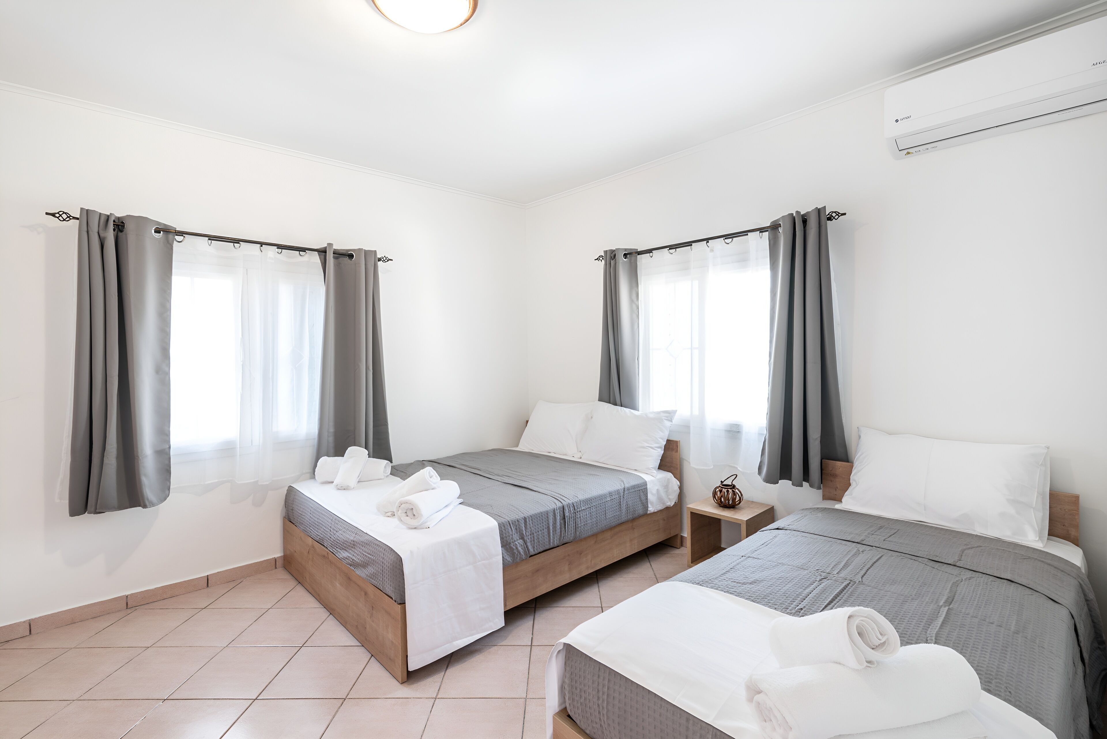 A hotel room with two beds, grey curtains, white bedding, and a tiled floor.