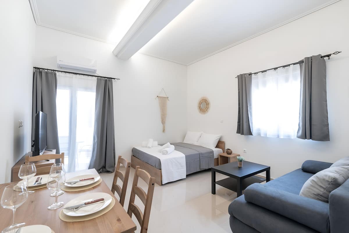 A modern, well-lit room with a bed, sofa, dining table set for two, and a small table with a plant.