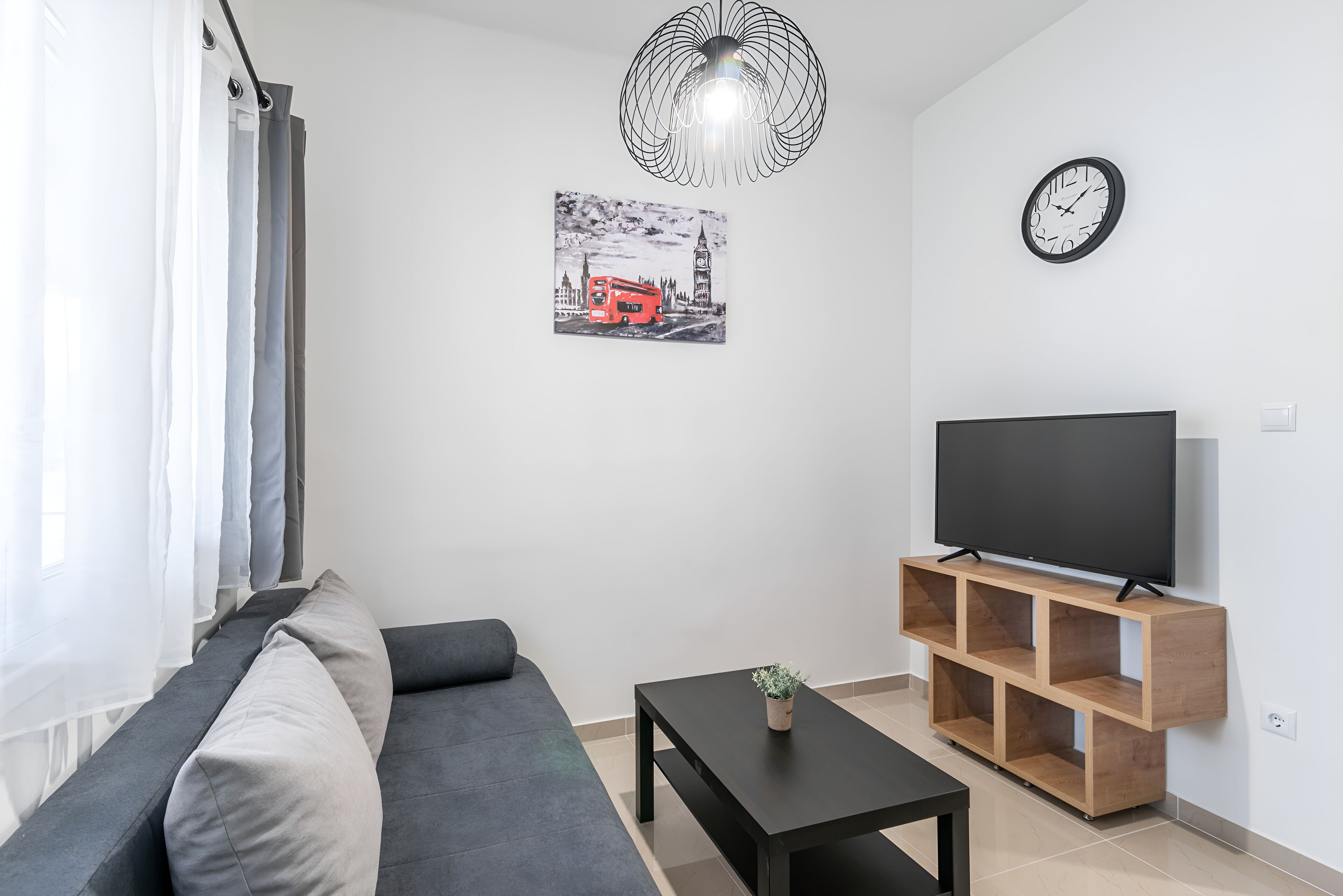 A modern living room with a grey sofa, a black coffee table, a TV on a wooden stand, a wall clock, and a black and white cityscape picture.