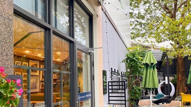 Un café con terraza, puerta de cristal y un pequeño jardín con macetas y sombrillas.