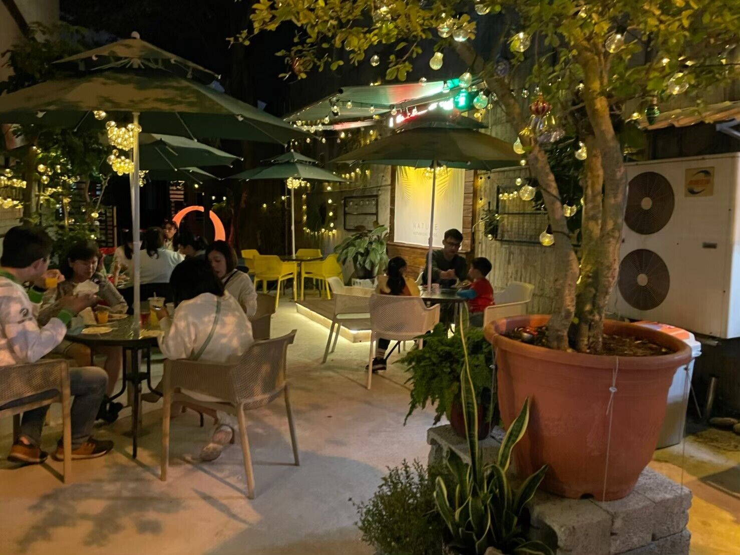 Un espace extérieur aménagé pour les repas, avec des tables et des chaises, entouré de plantes en pot et de parasols.