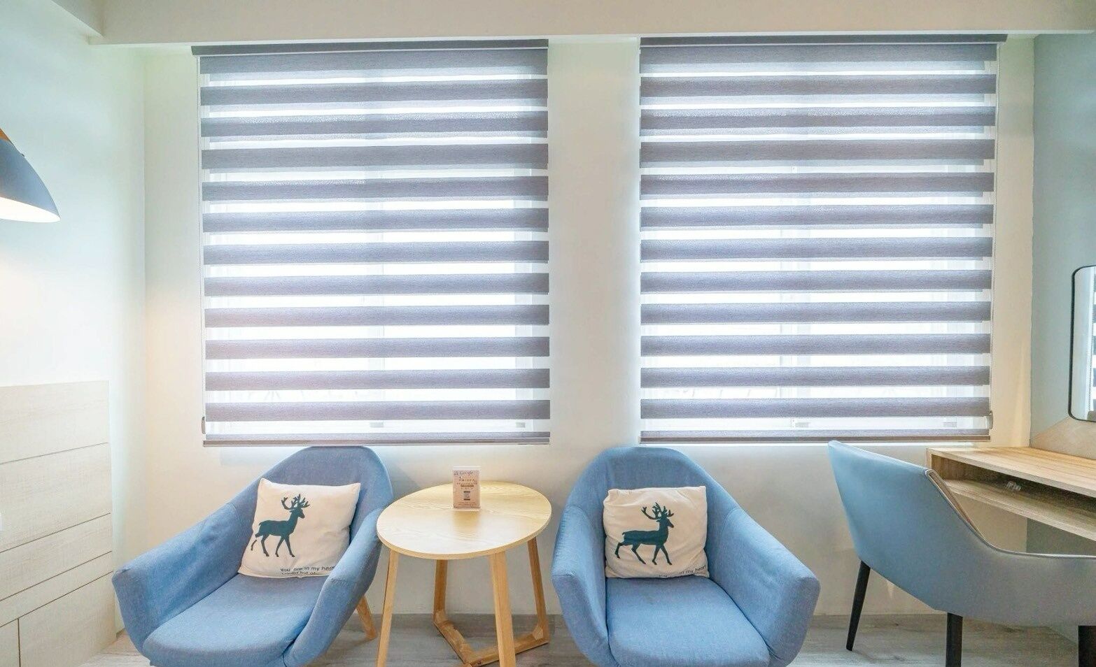 A room with two blue armchairs, a small round table, and two windows with horizontal blinds.