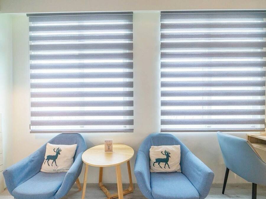 A room with two blue armchairs, a small round table, and two windows with horizontal blinds.