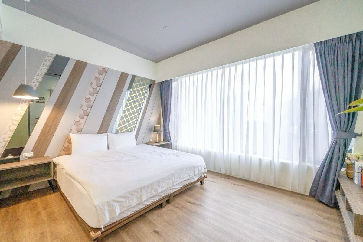 Une chambre à coucher moderne avec un grand lit, un plancher en bois et une vue sur l’extérieur à travers des rideaux transparents.