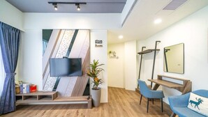 Una habitación de hotel moderna con un televisor de pantalla plana montado en una pared con diseño geométrico, piso de madera, un sillón azul, un escritorio con espejo y una planta en maceta.