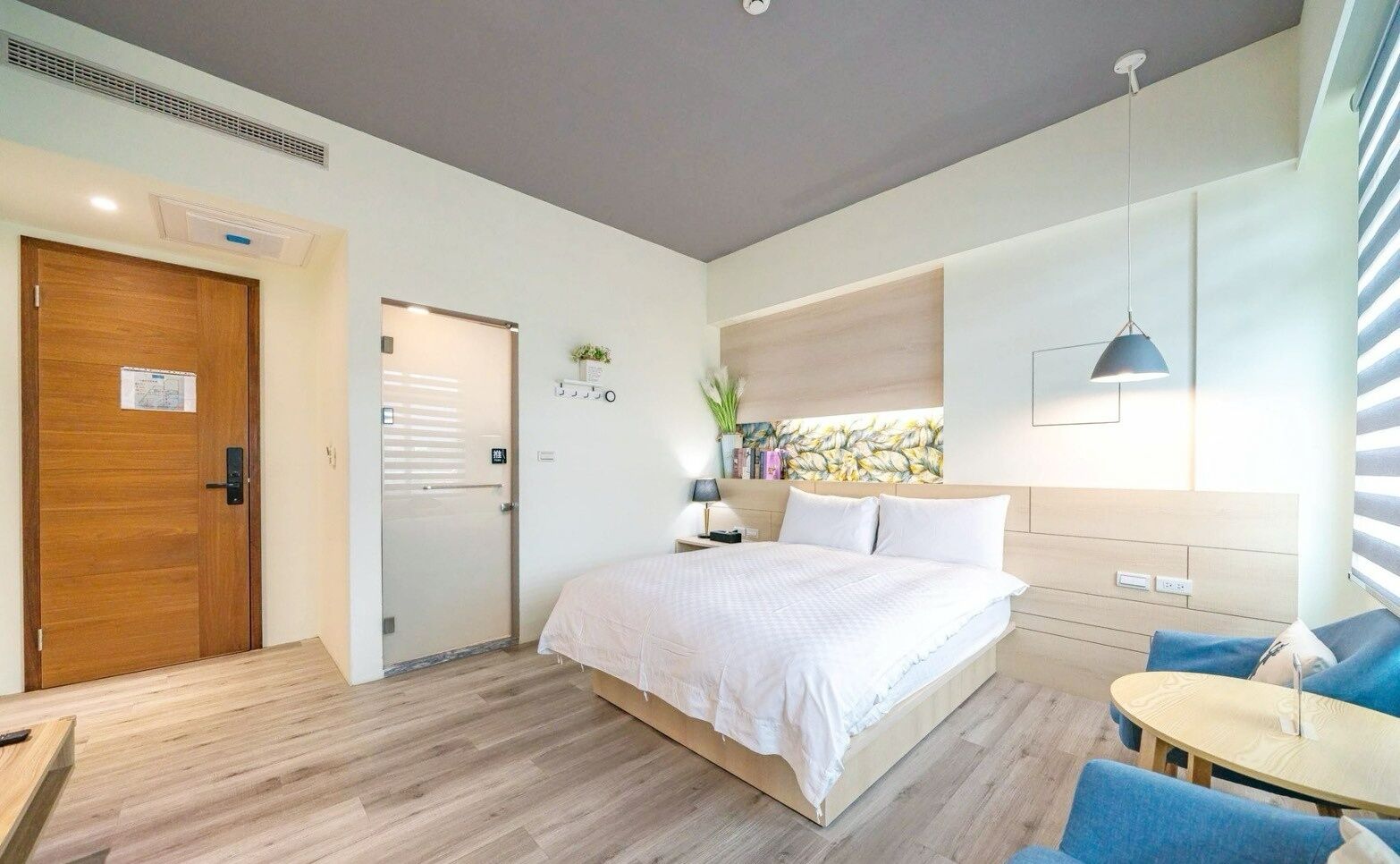 A modern hotel room with a wooden floor, a bed with white bedding, a small table with a lamp, and a view of the outside through large windows.