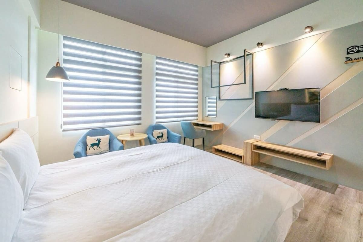 Une chambre d’hôtel moderne avec un grand lit, deux fauteuils bleus, une petite table, un téléviseur et un bureau avec un ordinateur.