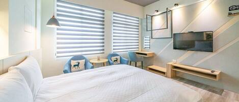 Una habitación de hotel moderna con una cama grande, dos sillas azules, una mesita, un televisor y un escritorio con una computadora.