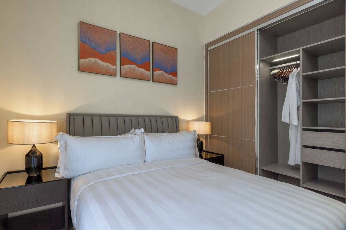 Une chambre d’hôtel avec un lit, deux lampes de chevet, une tête de lit grise et un dressing où sont suspendus des vêtements.