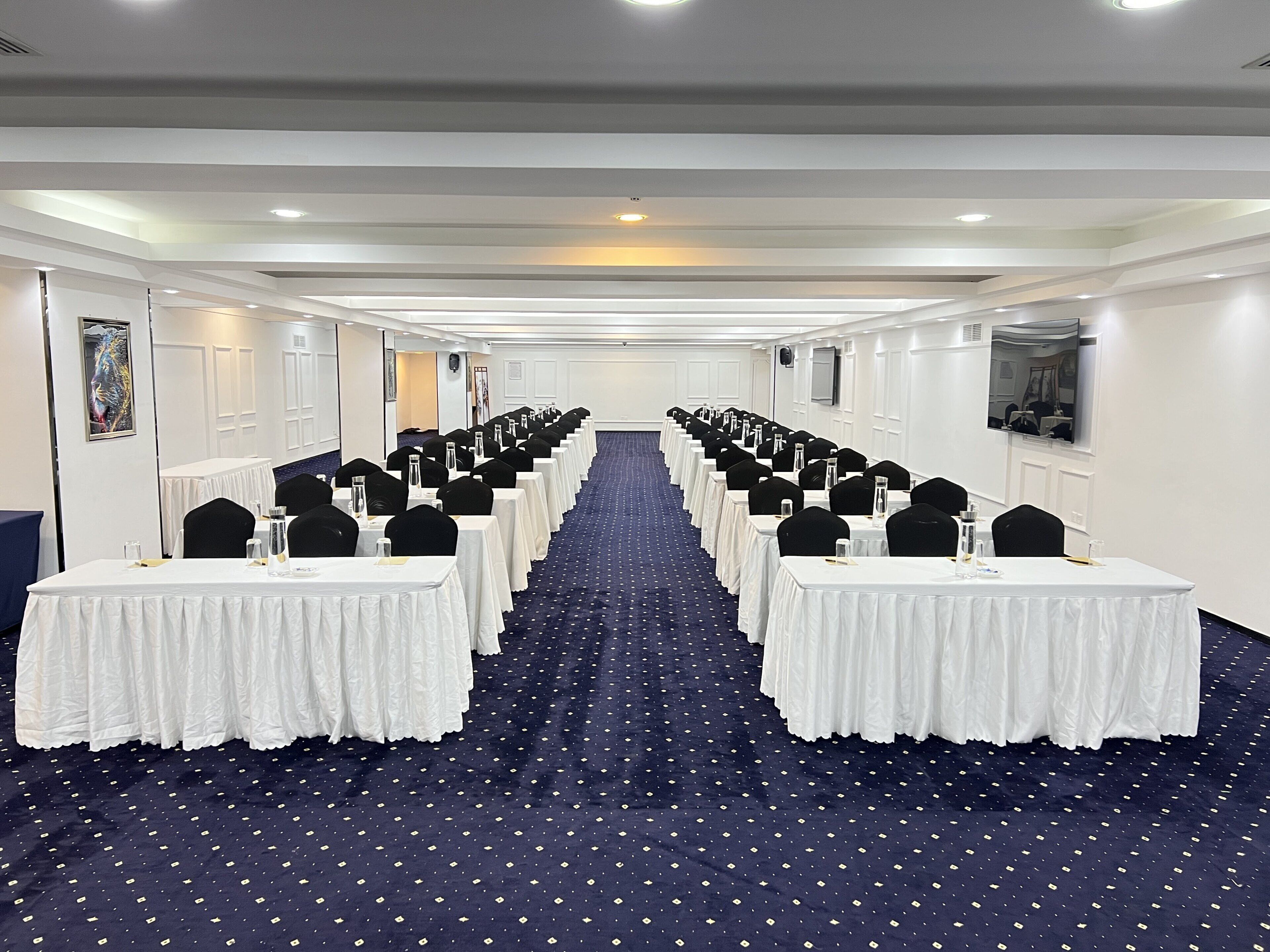A conference room with rows of tables and chairs, a patterned carpet, and a white ceiling with recessed lighting.