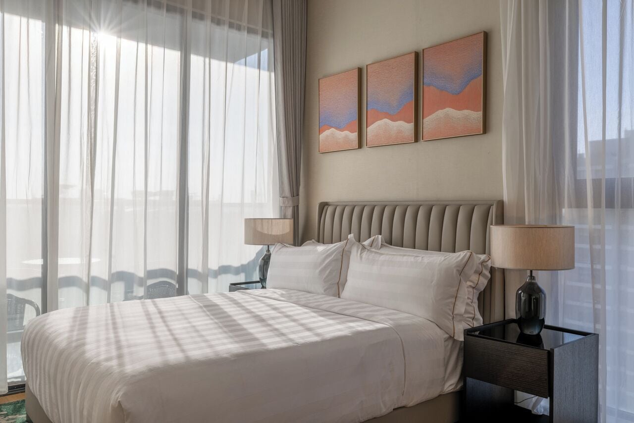 Una camera d'albergo con un letto grande, due comodini con lampade, vista attraverso tende leggere e tre quadri appesi al muro.