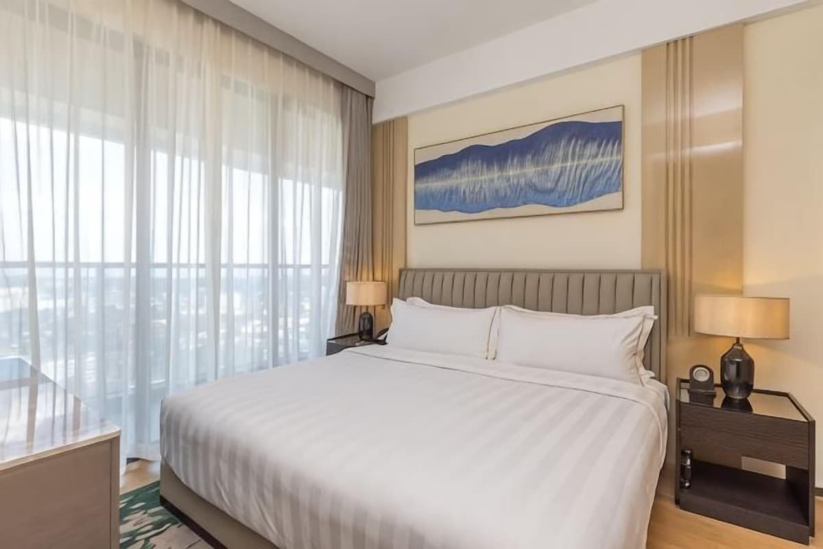Une chambre d’hôtel avec un grand lit, deux tables de chevet, une fenêtre avec des rideaux légers et un tableau au mur.