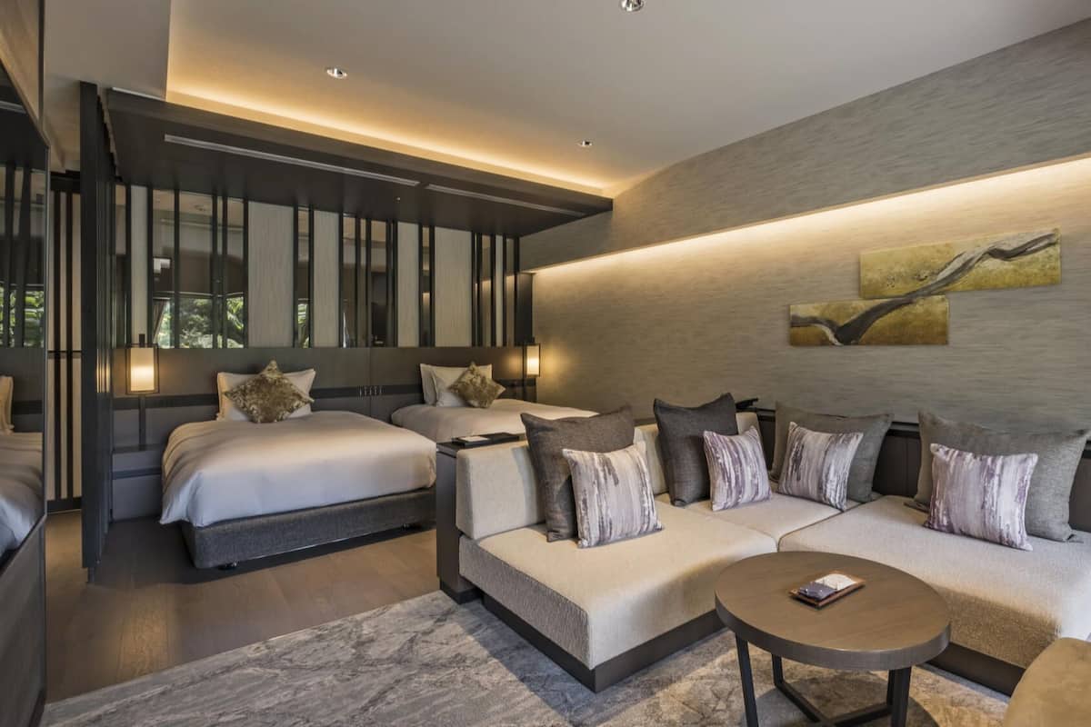 A modern hotel room with two beds, a sofa, a coffee table, and a large window.