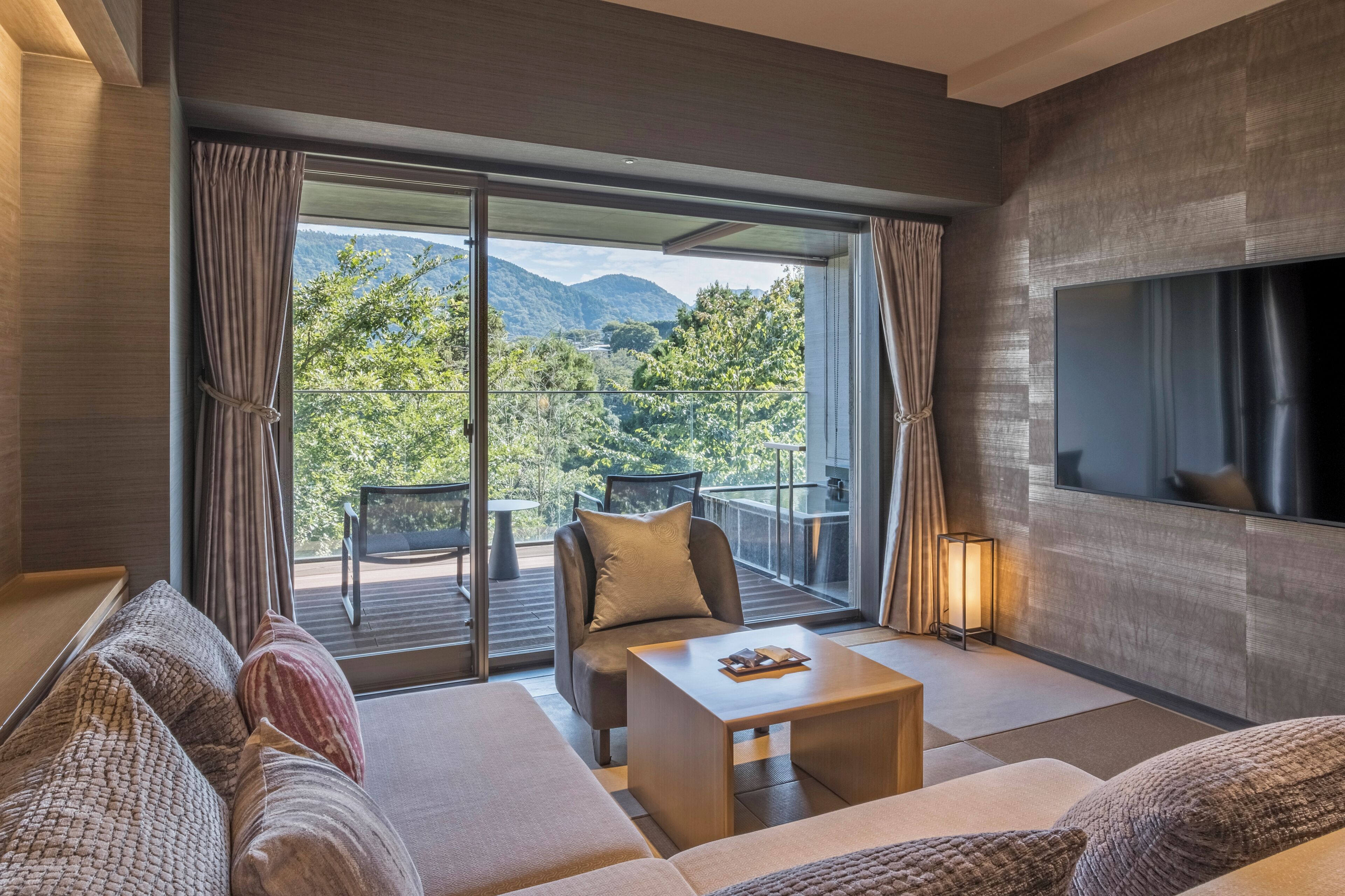 A modern hotel room with a sofa, a small table, a TV, and a balcony with a view of the mountains.