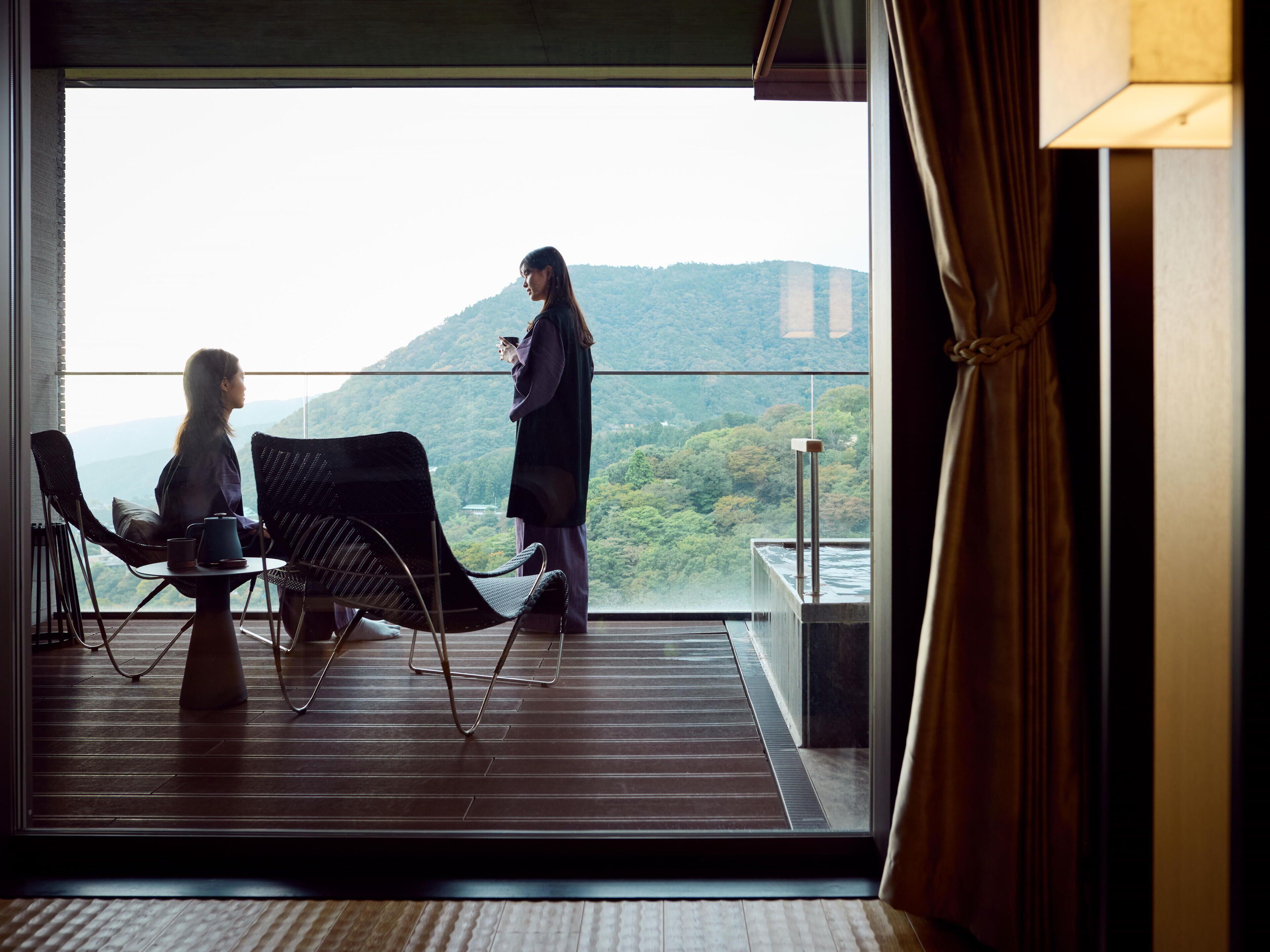 Un balcón con vistas a un paisaje montañoso, una mesa con sillas y una persona de pie.