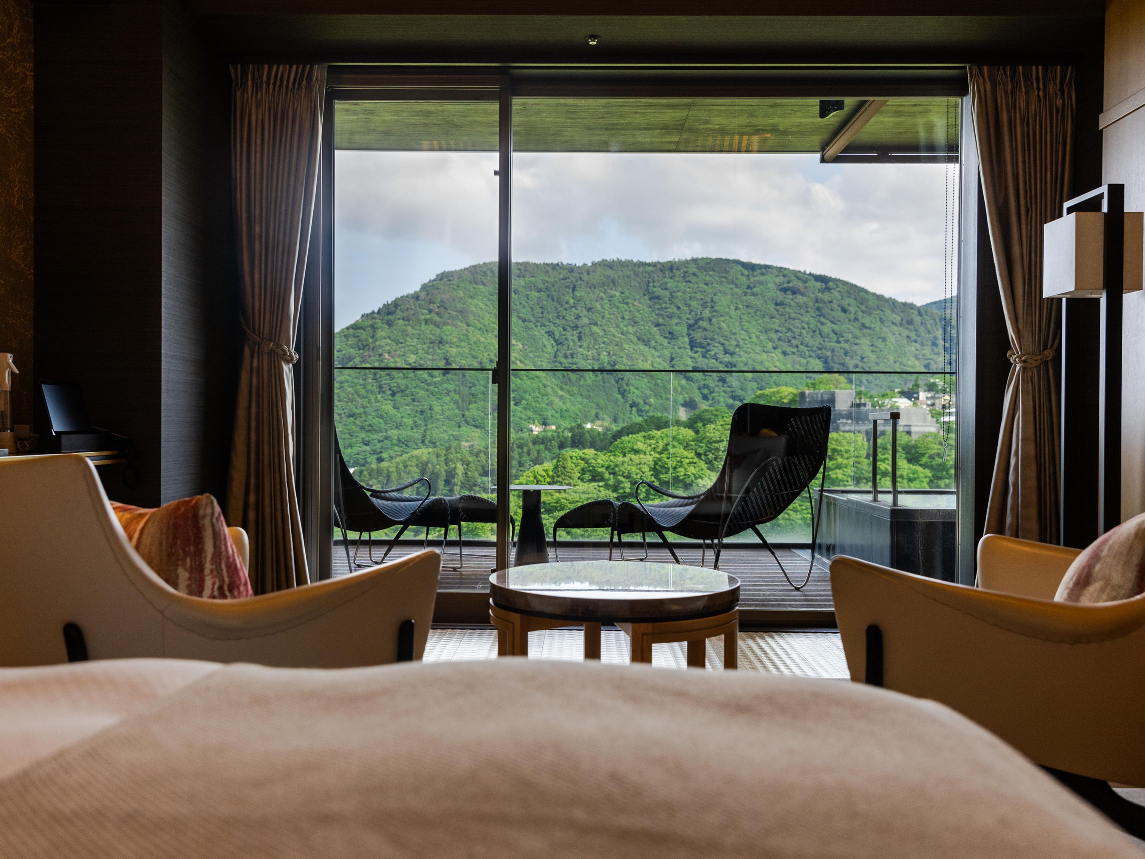Une chambre d’hôtel avec un grand lit, deux fauteuils, une petite table et une vue sur une montagne verdoyante.