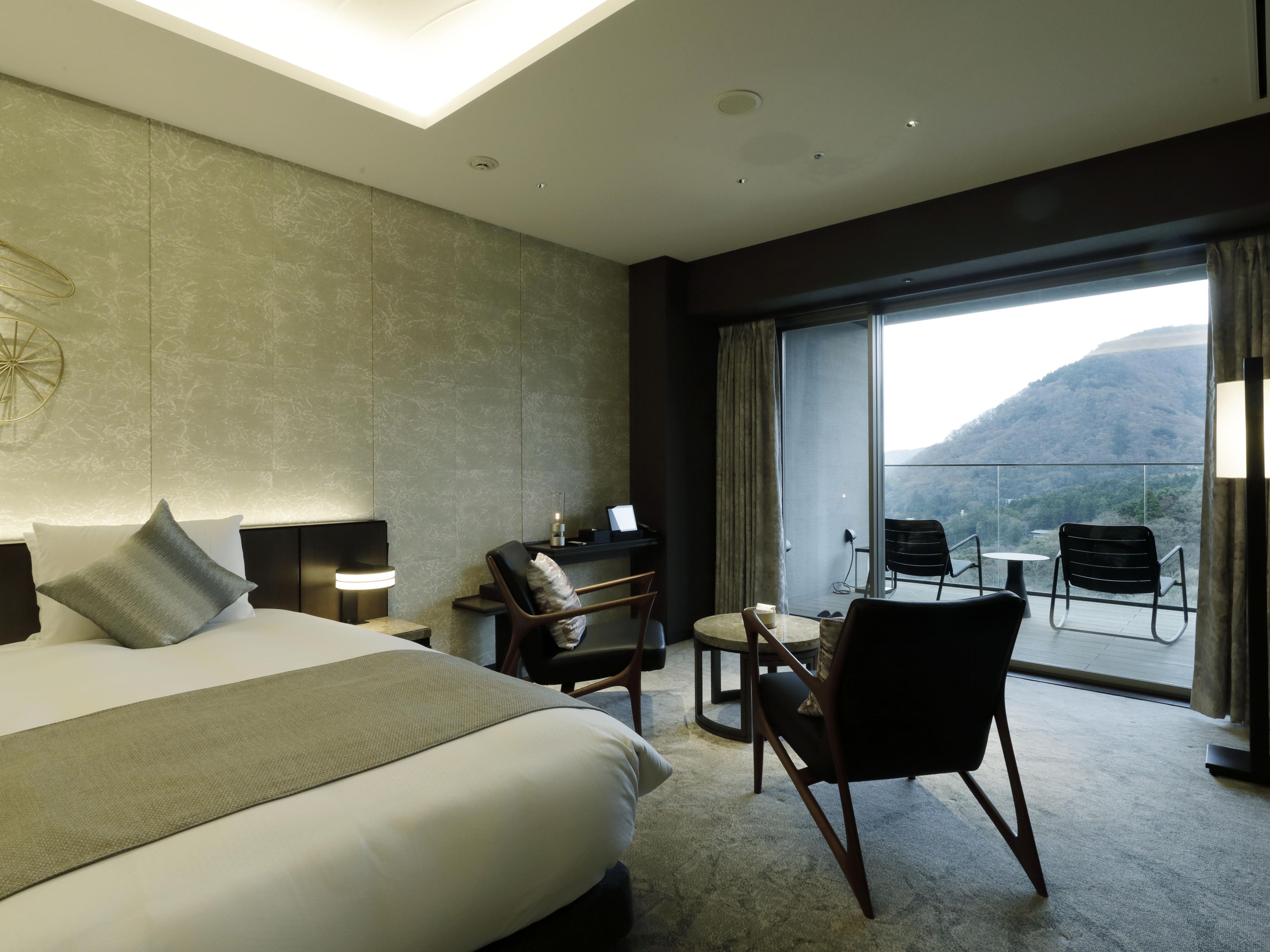 Ein Hotelzimmer mit einem Bett, einem Schreibtisch, zwei Stühlen, einem großen Fenster mit Bergblick und einer Deckenleuchte.