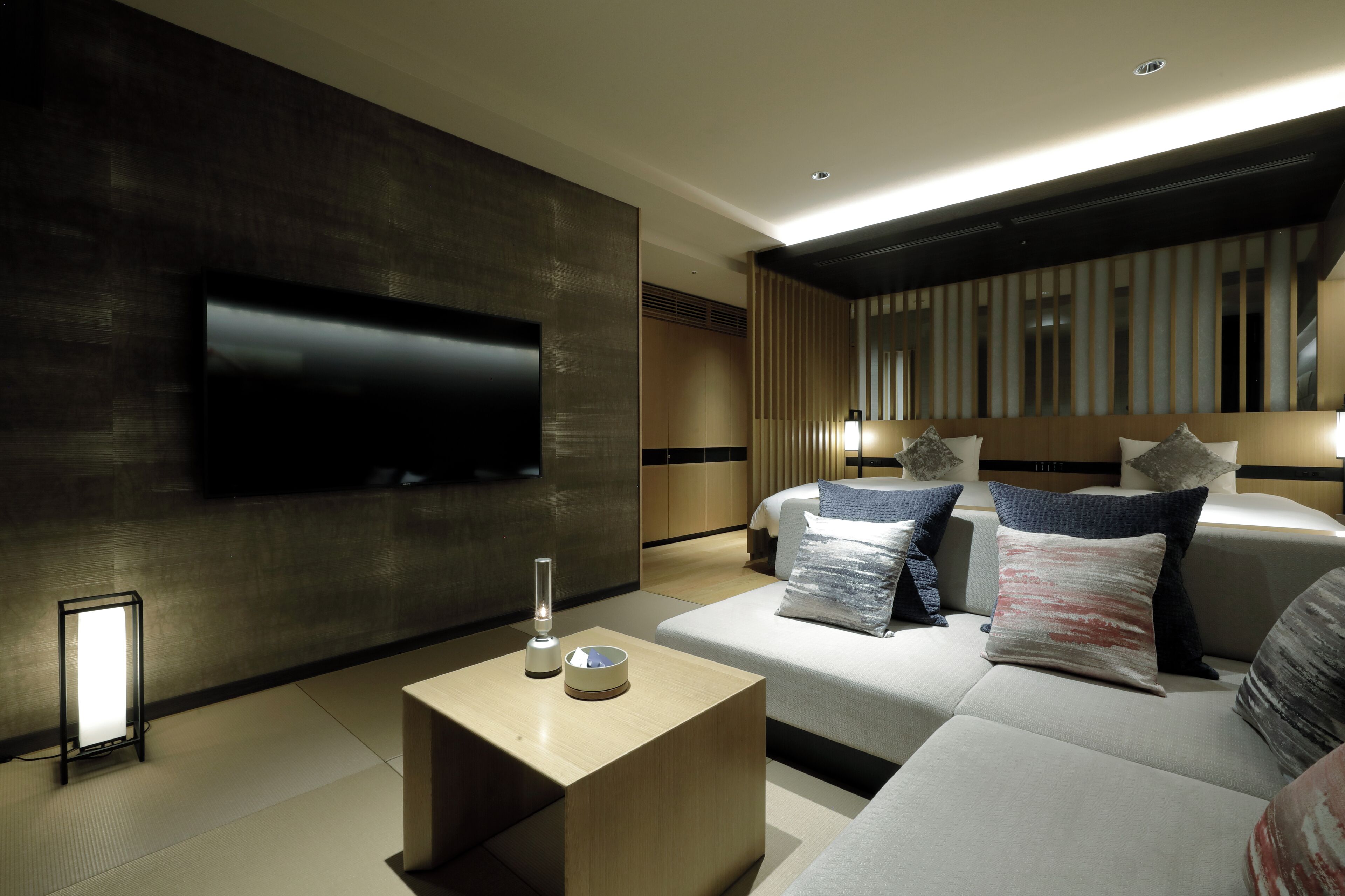 A modern hotel room with a large flat-screen TV mounted on the wall, a bedside table with a lamp, and a sofa with multiple pillows.
