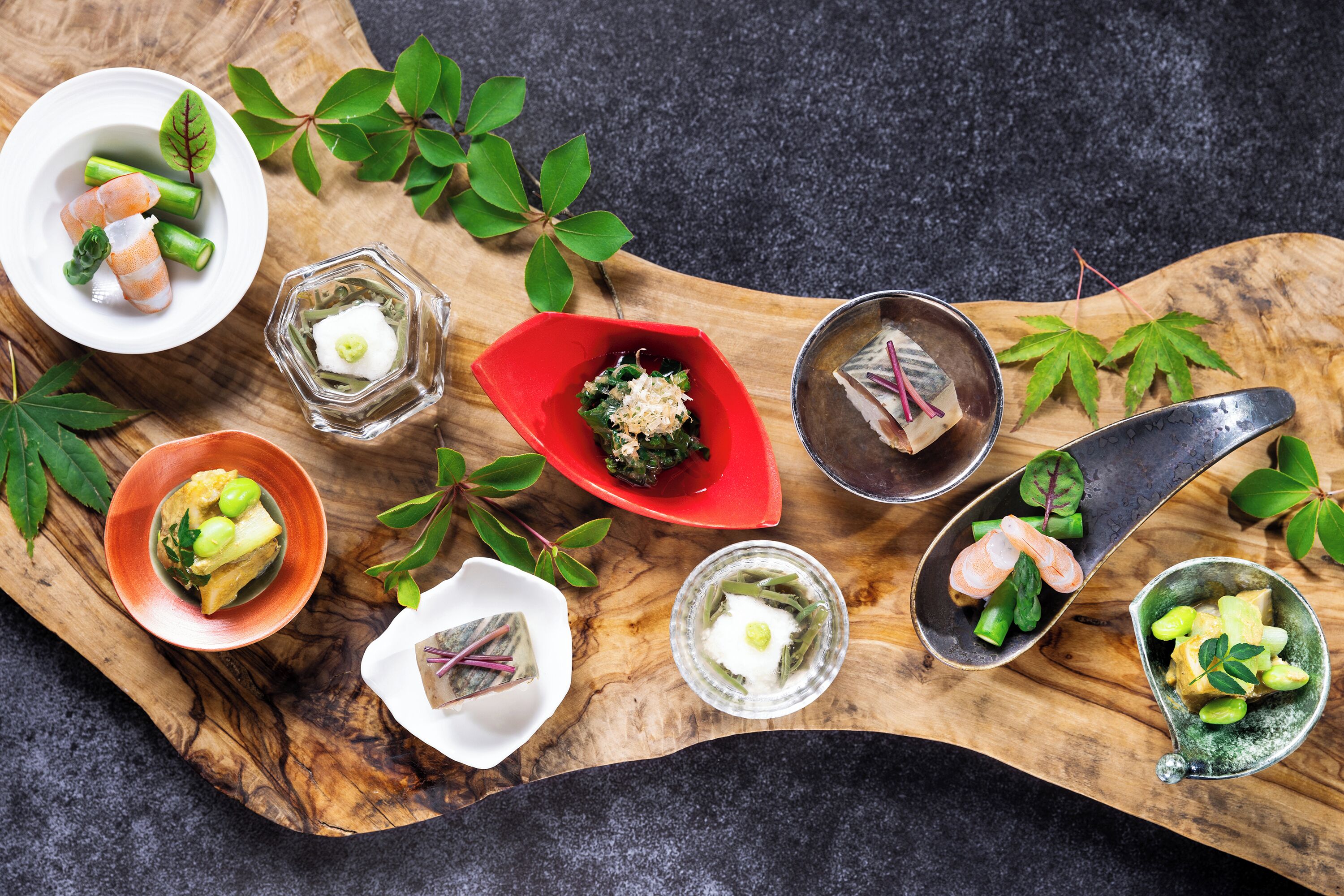 A variety of dishes arranged on a wooden board, including sushi, sashimi, and other Asian cuisine, with green leaves and small bowls as decorations.