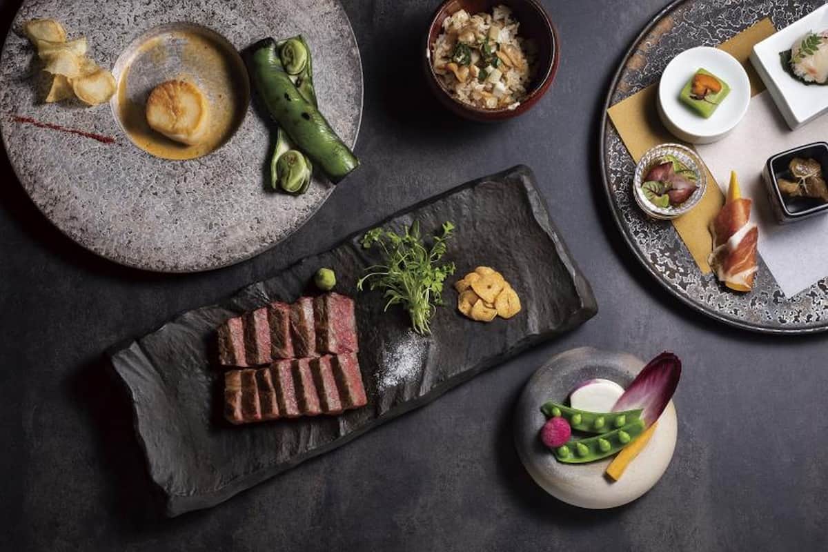 Ett urval av rätter, inklusive biff, grönsaker och sushi, presenterade på en mörk yta.