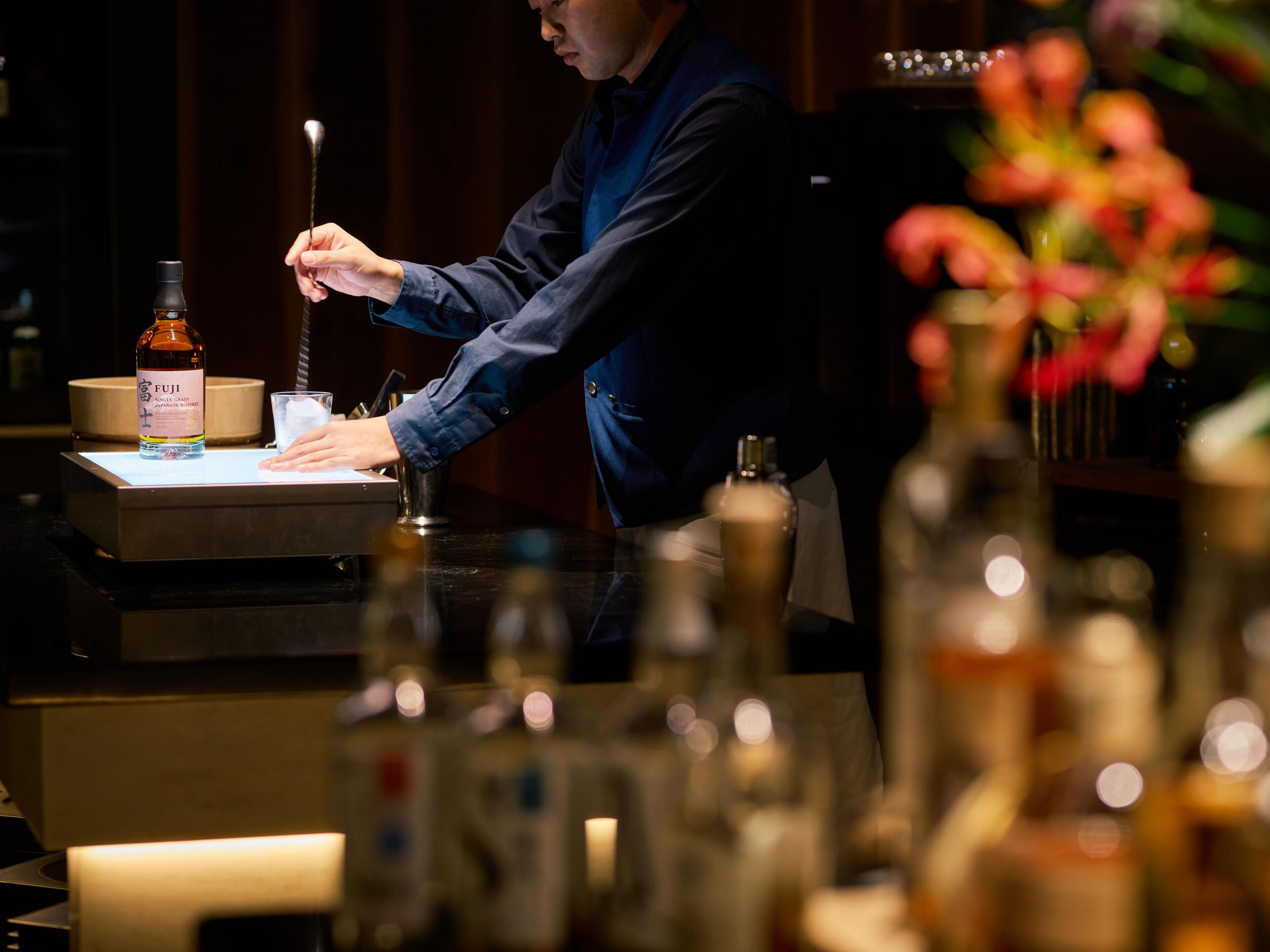 A bar setting with a person preparing a drink, a bottle labeled 'FUJI,' and various bar tools.