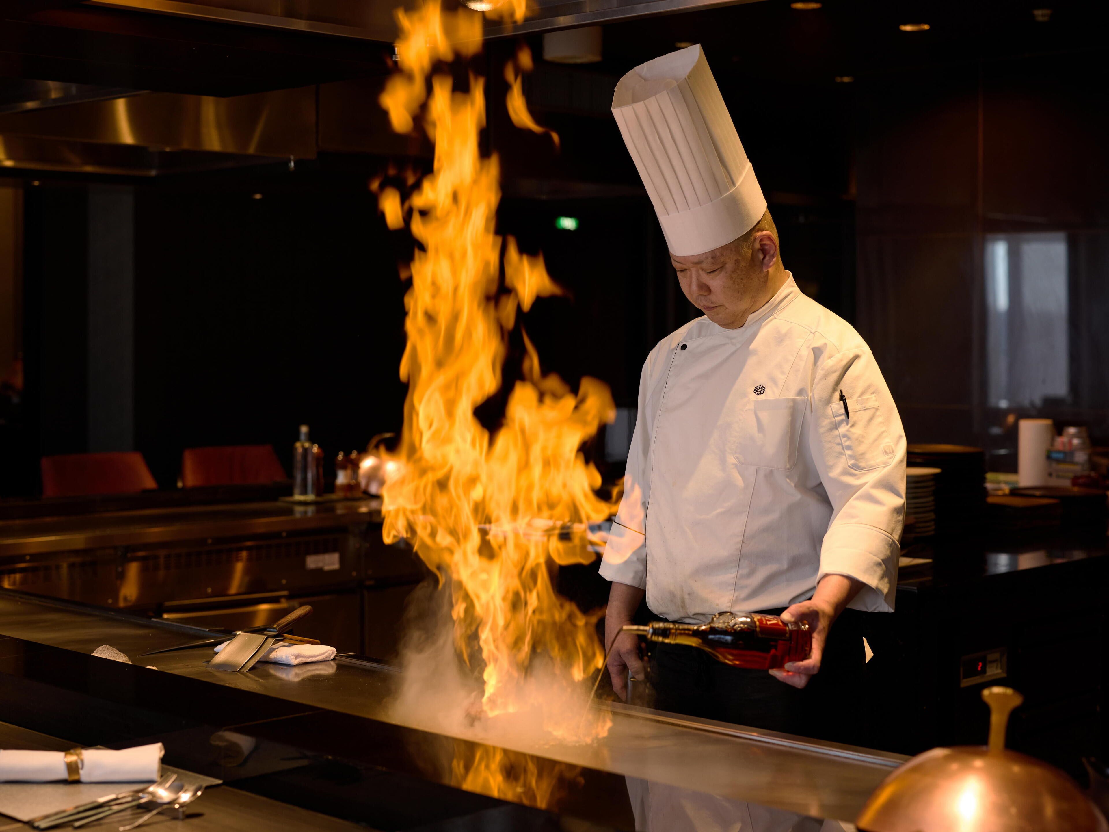 A chef performing a flambé with a bottle of liquor over a pan.