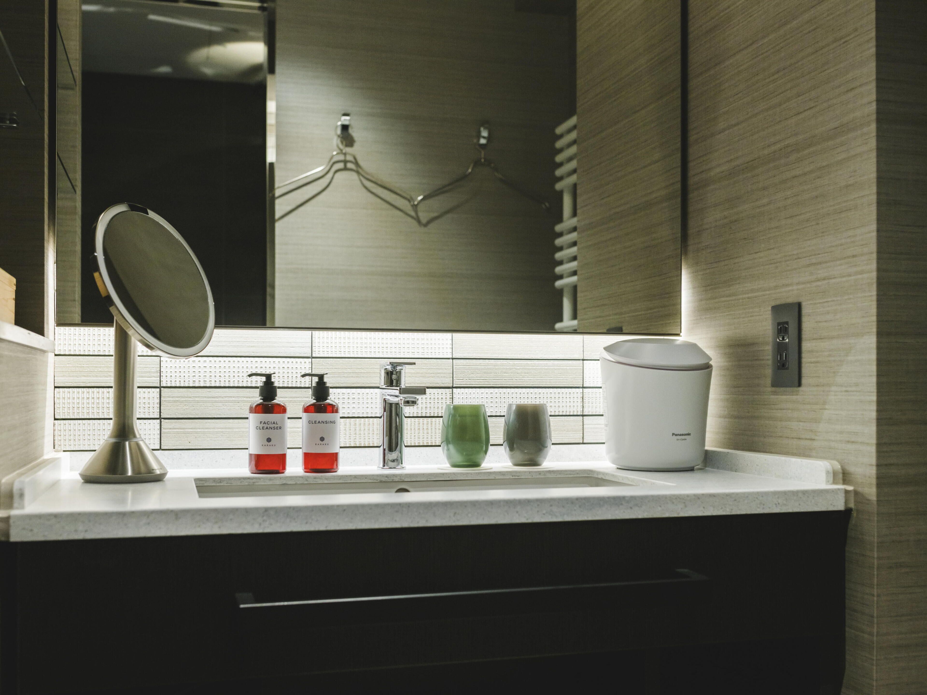 A modern bathroom with a sink, two bottles of facial cleanser, a green glass, and a white electric kettle.