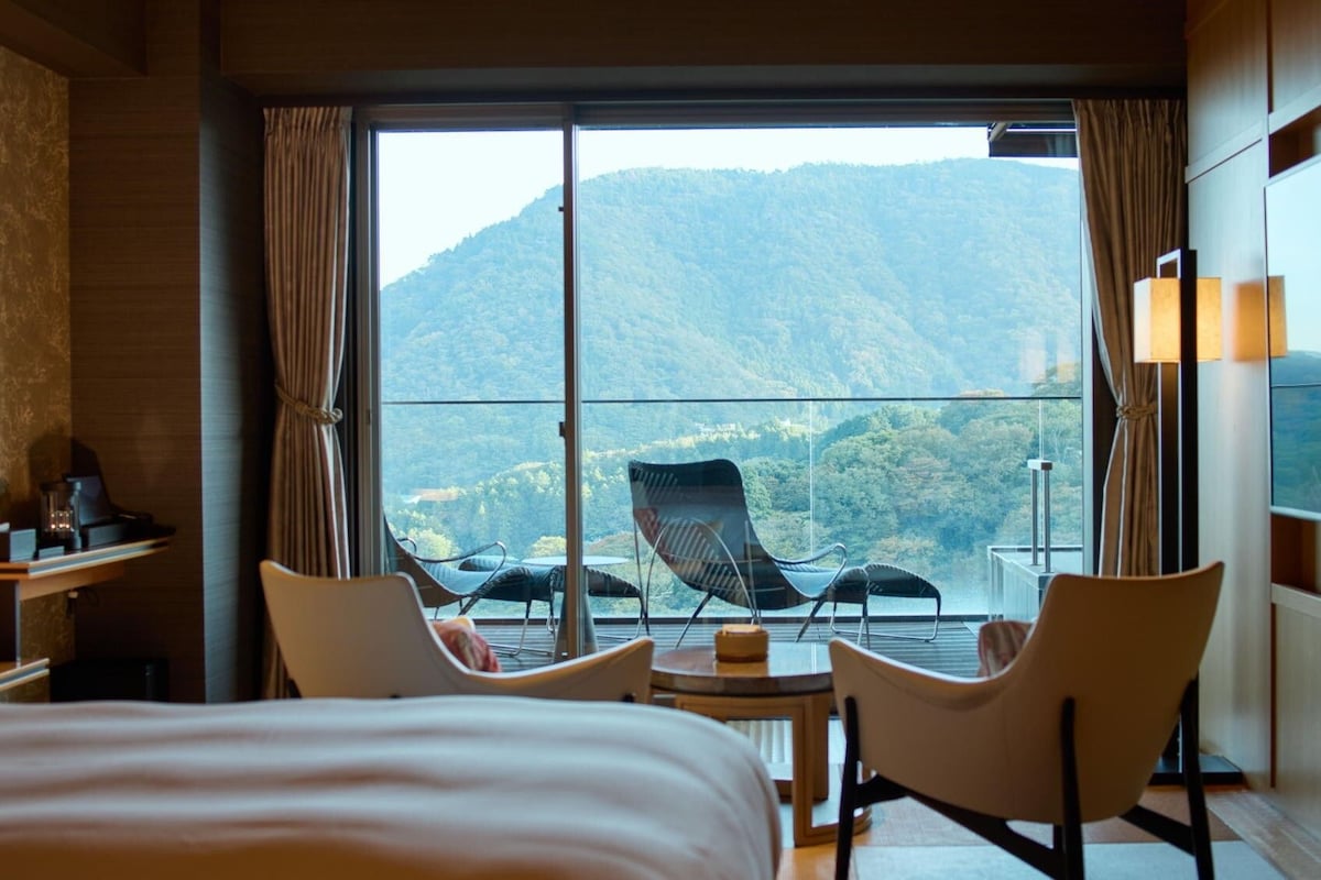 A hotel room with a large bed, two chairs, a small table, and a view of a mountainous landscape through a large window.