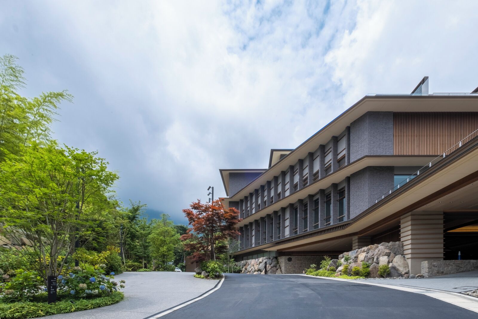Un bâtiment moderne doté d’un toit incurvé et de nombreuses fenêtres, entouré d’une végétation luxuriante et bordé d’une allée pavée.