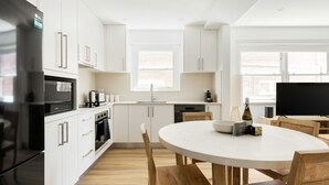 Une cuisine moderne avec des meubles blancs, un réfrigérateur noir, une table à manger avec des chaises et un téléviseur.