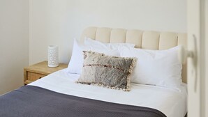 En pænt redt seng med en strukturmæssig pude og en glat pude, et lille natbord med en hvid lysestage, og en ensfarvet væg.