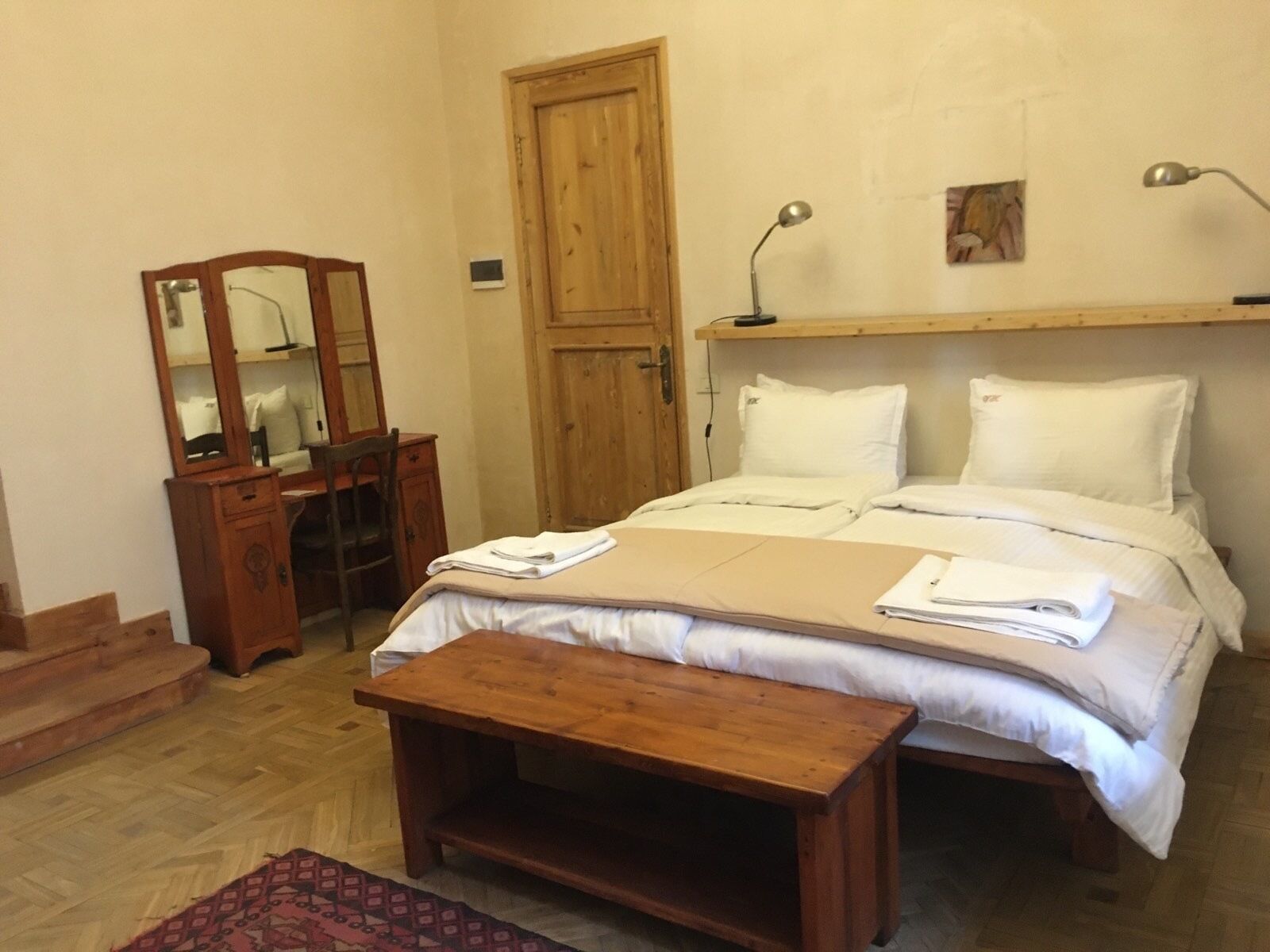 A bedroom with a wooden bed, a wooden desk with a mirror, a wooden bench, and a wooden door.
