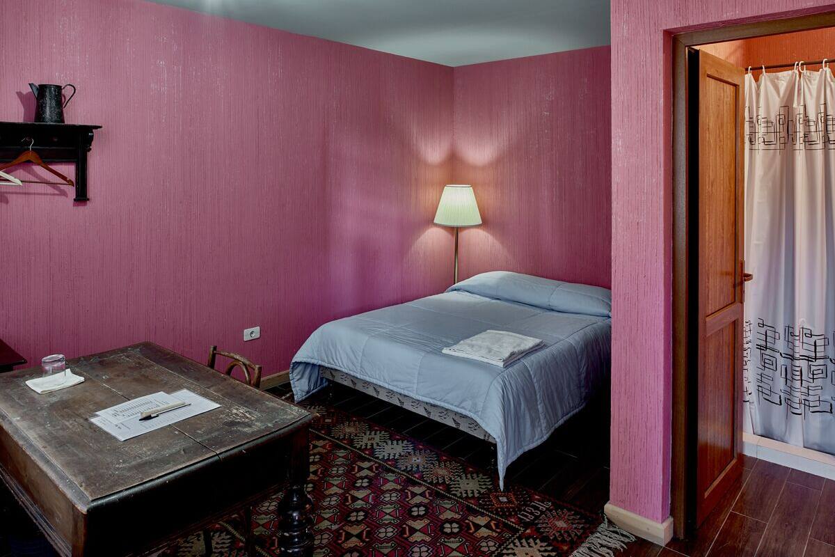 A room with pink walls, a bed with a blue bedspread, a wooden table with papers, a lamp, and a patterned rug.