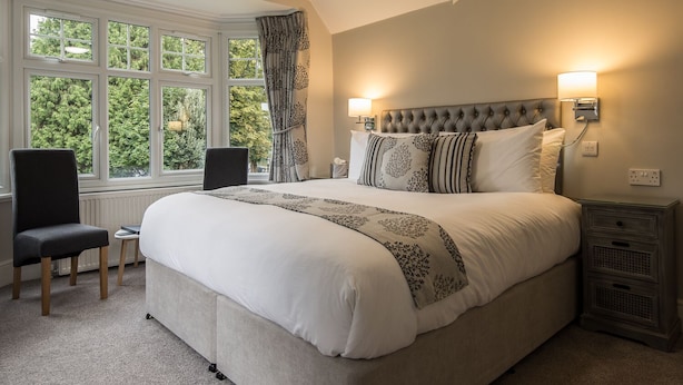 A bedroom with a large bed, two bedside lamps, a chair, and a view of trees through the window. - Maple Manor Hotel  (Crawley)