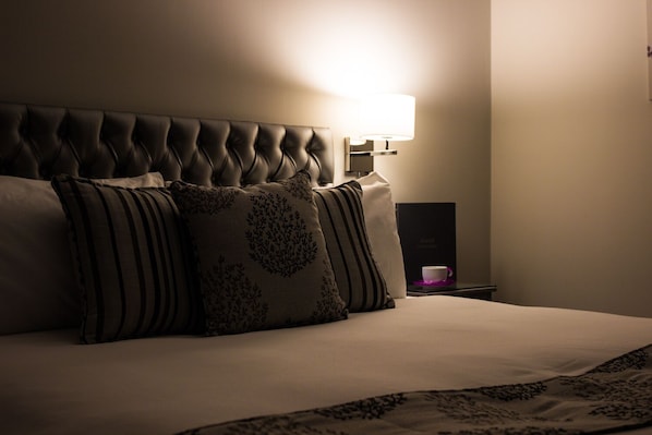 A neatly made bed with a tufted headboard, a bedside table with a lamp, and a cup on a saucer. - Maple Manor Hotel (Crawley)