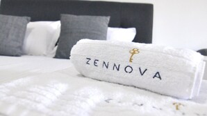 Un lit bien fait, avec une serviette blanche portant l’inscription « ZENNOVA » et un oreiller gris.