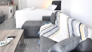 Une chambre d’hôtel moderne avec un canapé gris, une table basse en bois, un lit avec une literie blanche et un petit coin cuisine équipé d’un lave-linge.