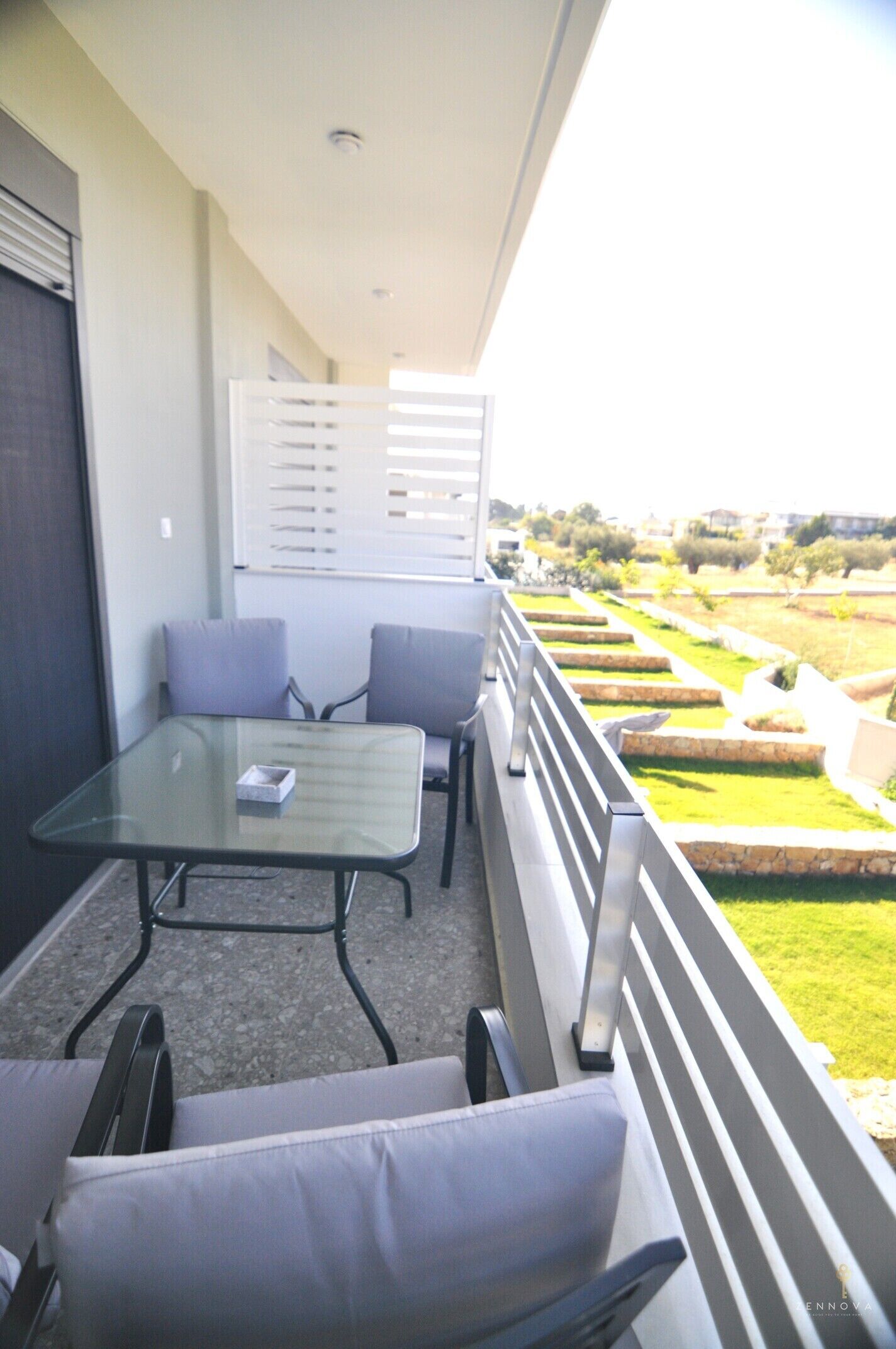A balcony with a glass table and two chairs, overlooking a landscaped garden.