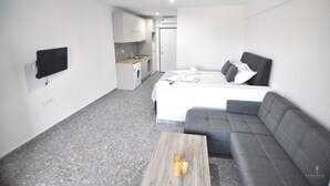 Une chambre d’hôtel compacte comprenant un lit, un canapé, une petite table, un téléviseur fixé au mur et, au fond, une kitchenette.